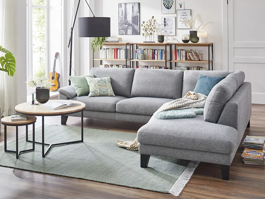 Moderne Wohnlandschaft in Grau mit Recamiere und Kissen, platziert vor bodentiefen Fenstern mit Blick ins Grüne. Stilvolles Sofa im hellen Wohnzimmer mit Teppich und Couchtisch als Inspiration für modernes Wohnen.